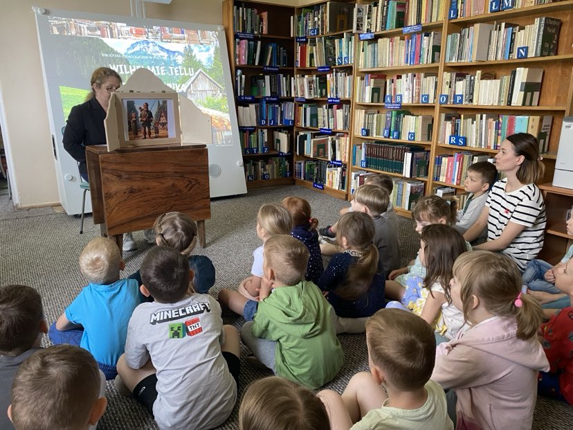 Na zdjęciu widzimy grupę dzieci siedzących na dywanie w bibliotece, skupionych na oglądaniu prezentacji i teatrzyku. Przed nimi ustawione jest urządzenie interaktywne, na którym wyświetlono ekran z napisem:
„Wilhelmie Tellu – traf w jabłko” – co sugeruje, że dzieci uczestniczą w zajęciach edukacyjno-ruchowych inspirowanych legendą o Wilhelmie Tellu.
Obok urządzenia stoi drewniana skrzynka teatrzyku kamishibai, w której umieszczona jest ilustracja przedstawiająca scenę z opowieści. Dzieci są zasłuchane i skupione, a obok nich siedzi nauczycielka, wspólnie z nimi uczestnicząc w wydarzeniu.
Całość odbywa się w przytulnej przestrzeni bibliotecznej, co pokazuje, jak nowoczesne technologie i tradycyjne formy przekazu (kamishibai) mogą być z powodzeniem łączone w pracy z dziećmi.