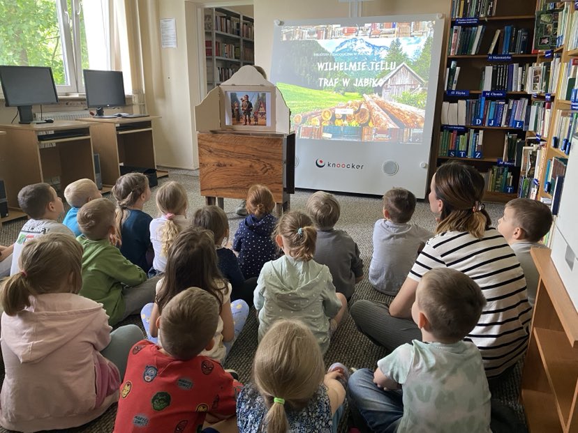 Na zdjęciu widzimy grupę dzieci siedzących na dywanie w bibliotece, skupionych na oglądaniu prezentacji i teatrzyku. Przed nimi ustawione jest urządzenie interaktywne, na którym wyświetlono ekran z napisem:
„Wilhelmie Tellu – traf w jabłko” – co sugeruje, że dzieci uczestniczą w zajęciach edukacyjno-ruchowych inspirowanych legendą o Wilhelmie Tellu.
Obok urządzenia stoi drewniana skrzynka teatrzyku kamishibai, w której umieszczona jest ilustracja przedstawiająca scenę z opowieści. Dzieci są zasłuchane i skupione, a obok nich siedzi nauczycielka, wspólnie z nimi uczestnicząc w wydarzeniu.
Całość odbywa się w przytulnej przestrzeni bibliotecznej, co pokazuje, jak nowoczesne technologie i tradycyjne formy przekazu (kamishibai) mogą być z powodzeniem łączone w pracy z dziećmi.