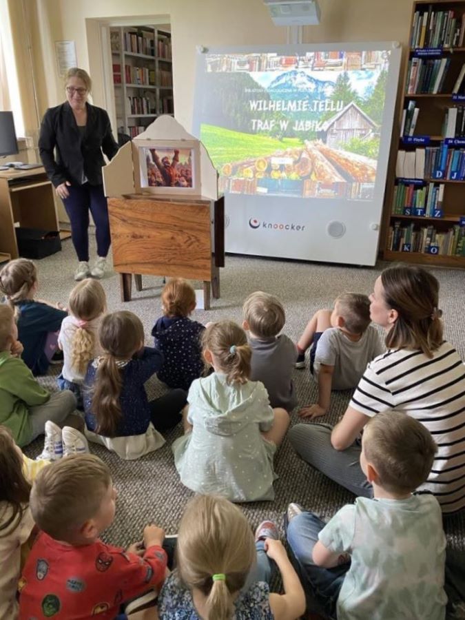 Na zdjęciu widzimy bibliotekarkę prowadzącą zajęcia dla dzieci w przestrzeni bibliotecznej. Grupa przedszkolaków siedzi na dywanie i z zainteresowaniem słucha opowieści. Za teatrzykiem znajduje się interaktywne urządzenie, na którym wyświetla się tytuł prezentowanej historii:
„Wilhelmie Tellu – traf w jabłko”.
Zajęcia są połączeniem tradycyjnej narracji wizualnej (kamishibai) oraz nowoczesnej technologii, co pozwala dzieciom lepiej zaangażować się w opowiadaną historię i aktywnie w niej uczestniczyć. Tło stanowią regały pełne książek, co tworzy ciepłą, edukacyjną atmosferę.