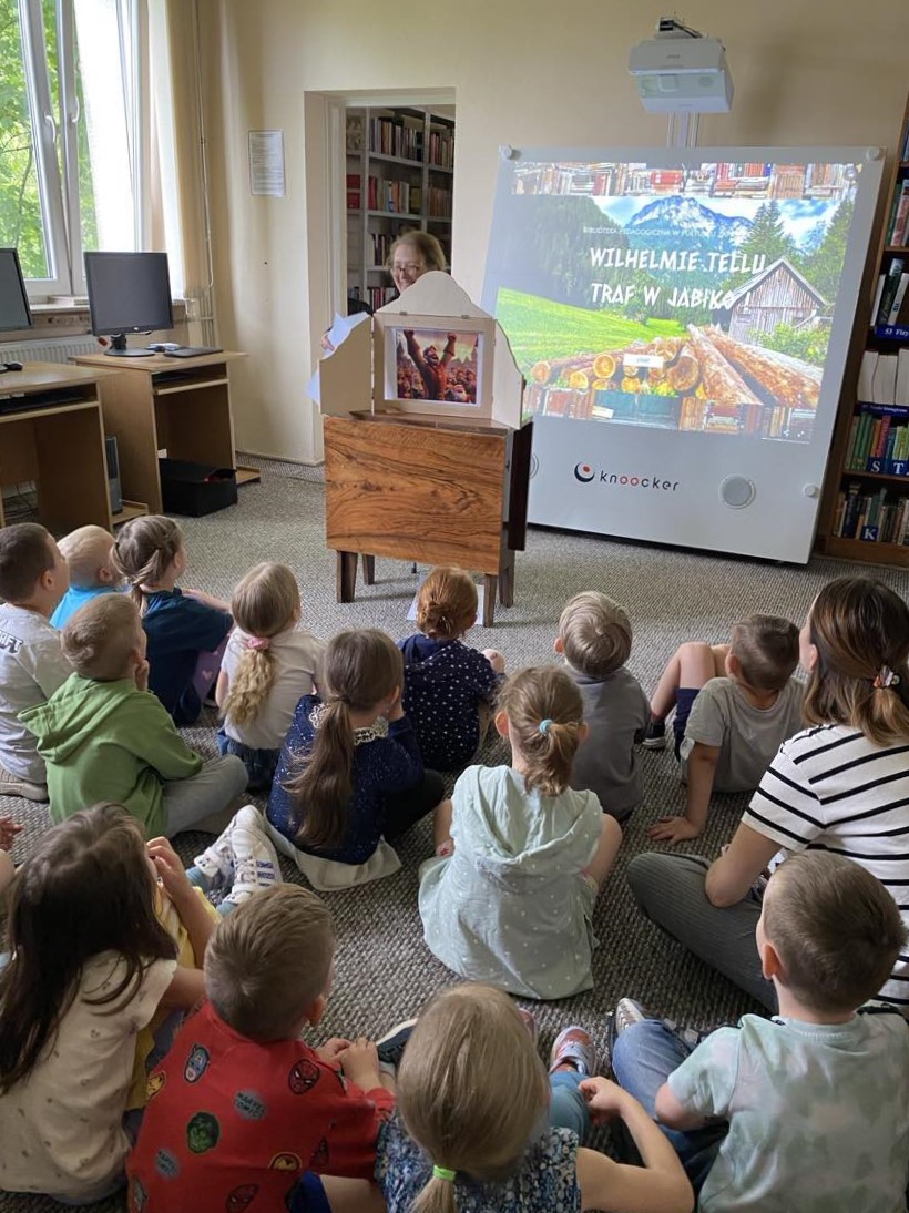 Na zdjęciu widzimy grupę dzieci siedzących na dywanie w bibliotece, skupionych na oglądaniu prezentacji i teatrzyku. Przed nimi ustawione jest urządzenie interaktywne, na którym wyświetlono ekran z napisem:
„Wilhelmie Tellu – traf w jabłko” – co sugeruje, że dzieci uczestniczą w zajęciach edukacyjno-ruchowych inspirowanych legendą o Wilhelmie Tellu.
Obok urządzenia stoi drewniana skrzynka teatrzyku kamishibai, w której umieszczona jest ilustracja przedstawiająca scenę z opowieści. Dzieci są zasłuchane i skupione, a obok nich siedzi nauczycielka, wspólnie z nimi uczestnicząc w wydarzeniu.
Całość odbywa się w przytulnej przestrzeni bibliotecznej, co pokazuje, jak nowoczesne technologie i tradycyjne formy przekazu (kamishibai) mogą być z powodzeniem łączone w pracy z dziećmi.