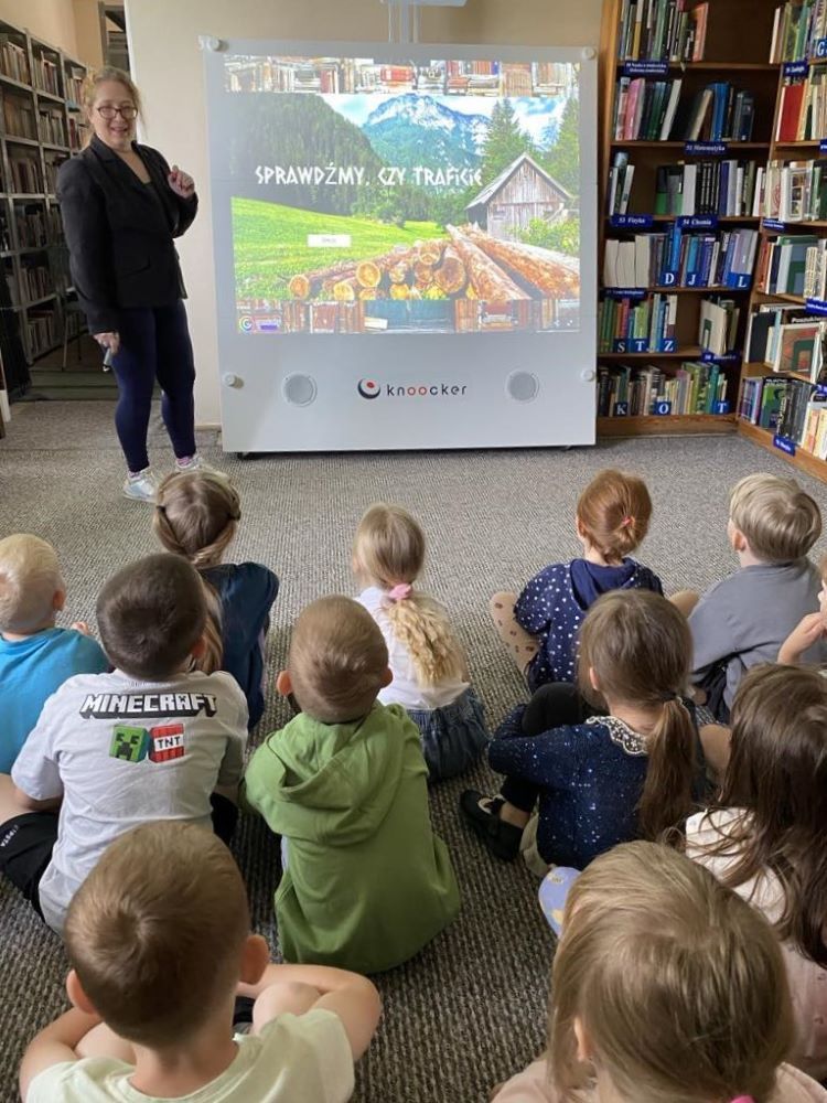 Na zdjęciu widzimy bibliotekarkę prezentującą siedzącym na dywanie dzieciom interaktywną grę edukacyjną. Na planszy wyświetla się napis: „Sprawdźmy czy, traficie?”, wyświetlany na urządzeniu Knoocker.
Dzieci uważnie śledzą przebieg gry – siedzą w rzędach na dywanie, skierowane w stronę ekranu. Pani prowadząca stoi z lewej strony planszy i tłumaczy zasady interaktywnej zabawy. Tło stanowią regały pełne książek, co wskazuje na przestrzeń biblioteczną, w której edukacja łączy się z zabawą i nowoczesną technologią.
To zdjęcie dokumentuje moment zaangażowania i skupienia dzieci podczas zajęć – świetny przykład nowoczesnej edukacji w bibliotece.