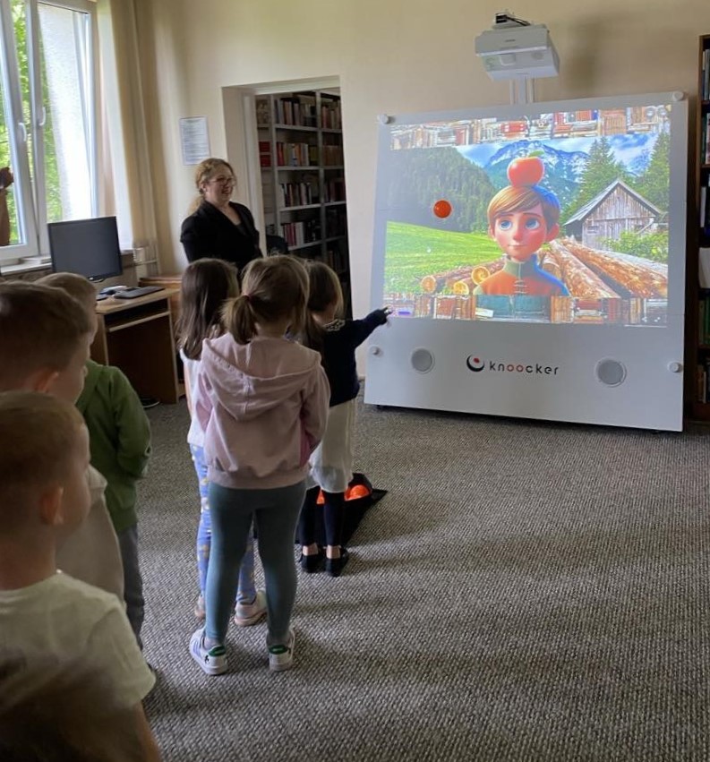 Na zdjęciu widać dzieci biorące udział w interaktywnej zabawie z wykorzystaniem urządzenia multimedialnego, które wyświetla kolorową animację z postacią na tle górskiego krajobrazu. Jedna z dziewczynek właśnie rzuca piłką w stronę ekranu, co wskazuje na element rywalizacji lub celowania w wybrane obiekty na ekranie. Obok dzieci stoi nauczycielka lub bibliotekarka, która koordynuje działania uczestników. W tle widoczne są regały z książkami i stanowisko komputerowe. Panuje atmosfera skupienia i radości – dzieci aktywnie uczestniczą w zabawie, która łączy elementy edukacyjne i ruchowe.