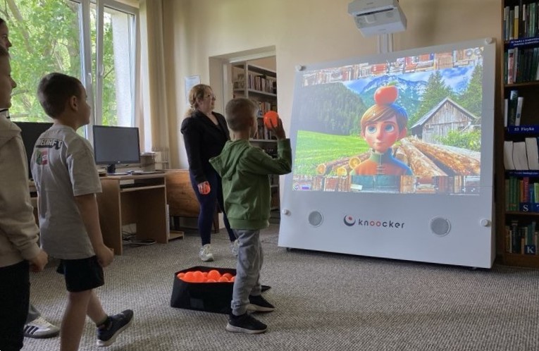 Na zdjęciu widać dzieci biorące udział w interaktywnej zabawie z wykorzystaniem urządzenia multimedialnego, które wyświetla kolorową animację z postacią na tle górskiego krajobrazu. Jeden z chłopców właśnie rzuca piłką w stronę ekranu, co wskazuje na element rywalizacji lub celowania w wybrane obiekty na ekranie. Obok dzieci stoi nauczycielka bibliotekarka, która koordynuje działania uczestników. W tle widoczne są regały z książkami i stanowisko komputerowe. Panuje atmosfera skupienia i radości – dzieci aktywnie uczestniczą w zabawie, która łączy elementy edukacyjne i ruchowe.