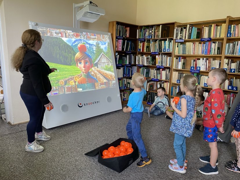 Na zdjęciu widać dzieci biorące udział w interaktywnej zabawie z wykorzystaniem urządzenia multimedialnego, które wyświetla kolorową animację z postacią na tle górskiego krajobrazu. Jeden z chłopców właśnie rzuca piłką w stronę ekranu, co wskazuje na element rywalizacji lub celowania w wybrane obiekty na ekranie. Obok dzieci stoi nauczycielka bibliotekarka, która koordynuje działania uczestników. W tle widoczne są regały z książkami i stanowisko komputerowe. Panuje atmosfera skupienia i radości – dzieci aktywnie uczestniczą w zabawie, która łączy elementy edukacyjne i ruchowe.