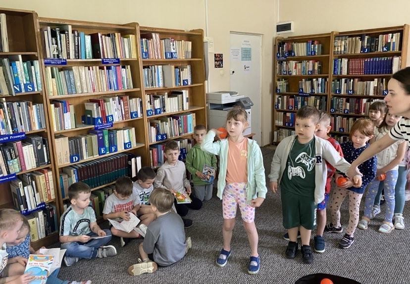 Na zdjęciu widzimy grupę dzieci uczestniczących w aktywności ruchowej w bibliotece. Dzieci ustawione są w kolejce, a jedno z nich – dziewczynka w różowych spodenkach – trzyma w ręku pomarańczową piłeczkę, przygotowując się do rzutu. Obok niej chłopiec i kolejne dzieci również trzymają podobne piłeczki, co sugeruje, że biorą udział w zabawie związanej z celowaniem w ramach gry „Wilhelmie Tellu, traf w jabłko”.
Po lewej stronie zdjęcia inne dzieci siedzą przy regale z książkami i czytają lub oglądają obrazki, co tworzy kontrast między zabawą ruchową a spokojniejszą aktywnością czytelniczą. W tle widać uporządkowane regały biblioteczne z oznaczeniami literowymi i urządzenie kserograficzne, co potwierdza, że akcja rozgrywa się w bibliotece.
Zajęcia łączące ruch i elementy edukacyjne w przestrzeni bibliotecznej rozwijają u dzieci nie tylko sprawność fizyczną, ale również kojarzenie literackich postaci i motywów – w tym przypadku legendarnego Wilhelma Tella.