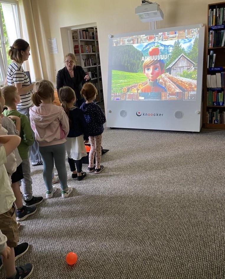 Na zdjęciu widać dzieci biorące udział w interaktywnej zabawie z wykorzystaniem urządzenia multimedialnego Knoocker, które wyświetla kolorową animację z postacią na tle górskiego krajobrazu. Jedna z dziewczynek właśnie rzuca piłką w stronę ekranu, co wskazuje na element rywalizacji lub celowania w wybrane obiekty na ekranie. Obok dzieci stoi nauczycielka lub bibliotekarka, która koordynuje działania uczestników. W tle widoczne są regały z książkami i stanowisko komputerowe. Panuje atmosfera skupienia i radości – dzieci aktywnie uczestniczą w zabawie, która prawdopodobnie łączy elementy edukacyjne i ruchowe.
