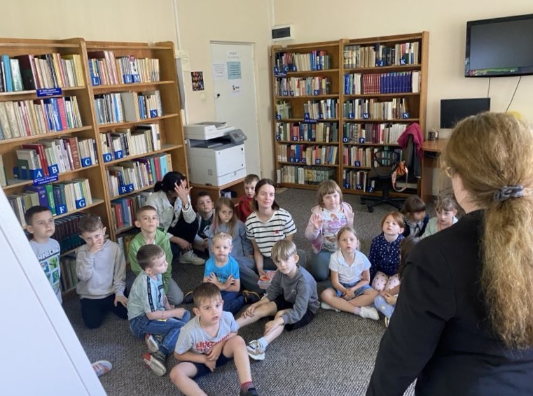 Na zdjęciu widzimy grupę dzieci siedzących na dywanie w bibliotece, skupionych na osobie prowadzącej (widocznej od tyłu), bibliotekarce. Dzieci wydają się uważne, niektóre trzymają w dłoniach kolorowe nagrody – co sugeruje, że biorą udział w podsumowaniu zajęć lub rozdaniu upominków po zakończonej aktywności.
W tle znajdują się regały z książkami, wyraźnie oznaczone literami alfabetu, co ułatwia poruszanie się po księgozbiorze. Widać również kserokopiarkę, stanowisko komputerowe i telewizor zawieszony na ścianie – typowe wyposażenie współczesnej biblioteki.
Atmosfera na zdjęciu jest przyjazna i sprzyjająca nauce oraz zabawie. Zdjęcie dokumentuje ważny moment edukacyjnego spotkania, który może być podsumowaniem lub pożegnaniem z uczestnikami.
