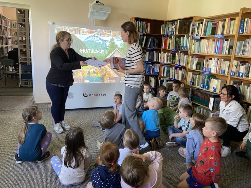 Na zdjęciu widzimy zakończenie spotkania edukacyjnego z dziećmi w bibliotece. W centrum kadru znajduje się bibliotekarka, która wręcza nauczycielce podziękowanie. W tle na ekranie wyświetlony jest napis „GRATULACJE!” oraz grafika z gry edukacyjnej „Wilhelmie Tellu, traf w jabłko”, co wskazuje na zakończenie zabawy, warsztatu.
Dzieci siedzące na dywanie obserwują wydarzenie, a ich postawa sugeruje zainteresowanie i skupienie. W tle widoczne są regały z książkami i uporządkowany księgozbiór, a także nauczycielka wspierająca grupę, siedząca z dziećmi.
Zdjęcie dokumentuje finał ciekawego spotkania łączącego literaturę, technologię i aktywność ruchową, co widać po zaangażowaniu dzieci oraz pozytywnej atmosferze wydarzenia.