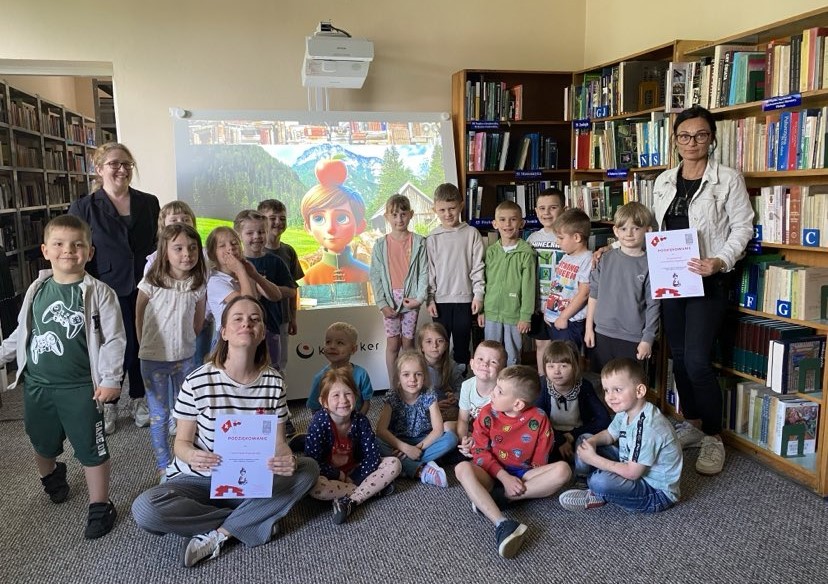 Na zdjęciu widzimy grupę dzieci wraz z opiekunkami oraz bibliotekarką, pozujących wspólnie na tle interaktywnego urządzenia , które wyświetla kolorową postać syna Wilhelma Tella z bajkowego krajobrazu. Zdjęcie zostało wykonane w bibliotece, o czym świadczą widoczne w tle regały pełne książek. Kilka osób trzyma w rękach dyplomy z podziękowaniami ze spotkania. Dzieci są uśmiechnięte, zrelaksowane i najwyraźniej zadowolone z udziału w wydarzeniu. Atmosfera jest bardzo pozytywna i integracyjna.