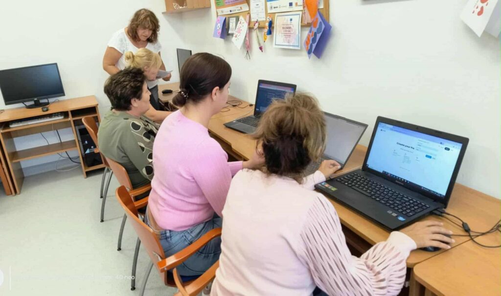 W sali komputerowej cztery kobiety siedzą przy biurkach, pracując na laptopach. Za nimi stoi prowadząca, która pomaga osobie przy stanowisku. Na ścianie nad biurkami wiszą certyfikaty i dyplomy, a przestrzeń jest uporządkowana i sprzyja nauce. Całość sugeruje szkolenie lub warsztaty z zakresu obsługi komputera w przyjaznym, edukacyjnym środowisku.