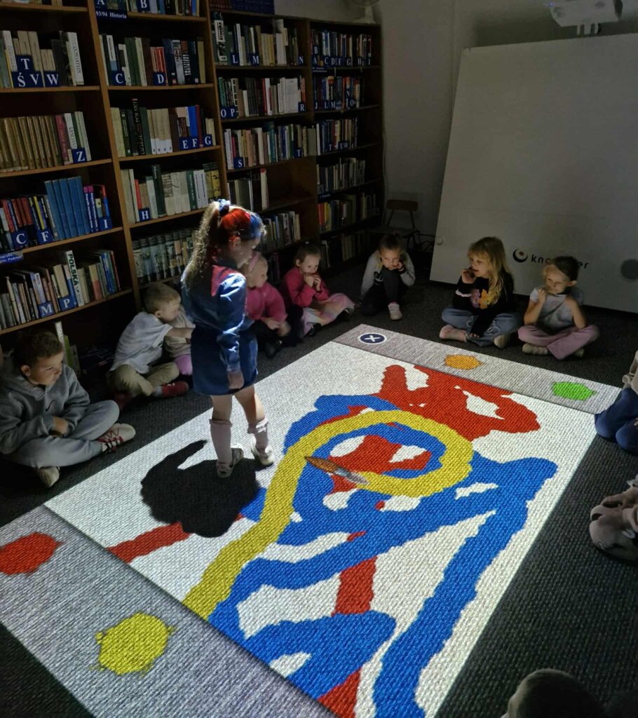 Na zdjęciu grupa dzieci siedzi na podłodze w bibliotece, a jedna dziewczynka stoi, wchodząc w interakcję z dużą, interaktywną projekcją na dywanie. Projekcja wyświetla obraz przypominający aplikację do rysowania, z dużymi, kolorowymi pociągnięciami pędzla w odcieniach niebieskim, żółtym i czerwonym. W tle widać regały pełne książek.