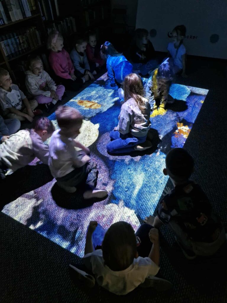 Na zdjęciu grupa dzieci siedzi na podłodze w bibliotece, a na dywanie wyświetlana jest duża, kolorowa projekcja. Dzieci wchodzą z nią w interakcję, a światło z projektora tworzy na nich barwne plamy. W tle widać regały pełne książek.
