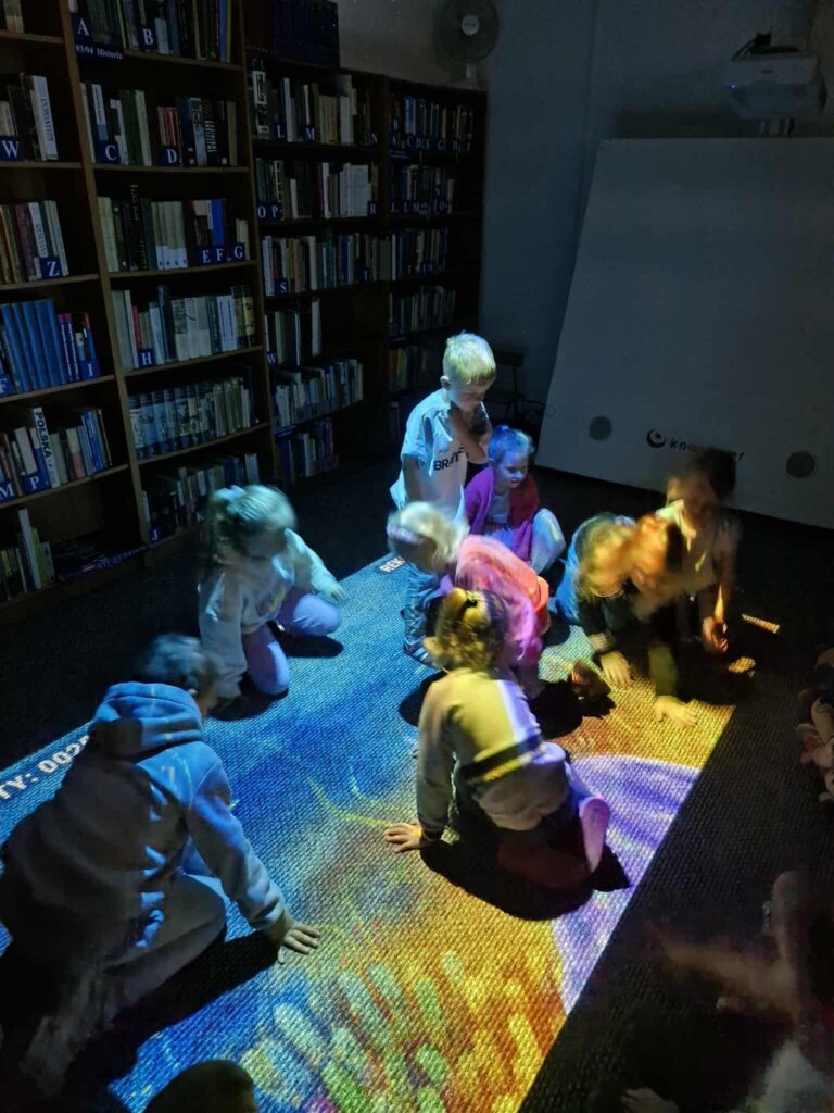 Na zdjęciu grupa dzieci siedzi na podłodze w bibliotece, a na dywanie wyświetlana jest duża, kolorowa projekcja. Dzieci wchodzą z nią w interakcję, a światło z projektora tworzy na nich barwne plamy. W tle widać regały pełne książek.