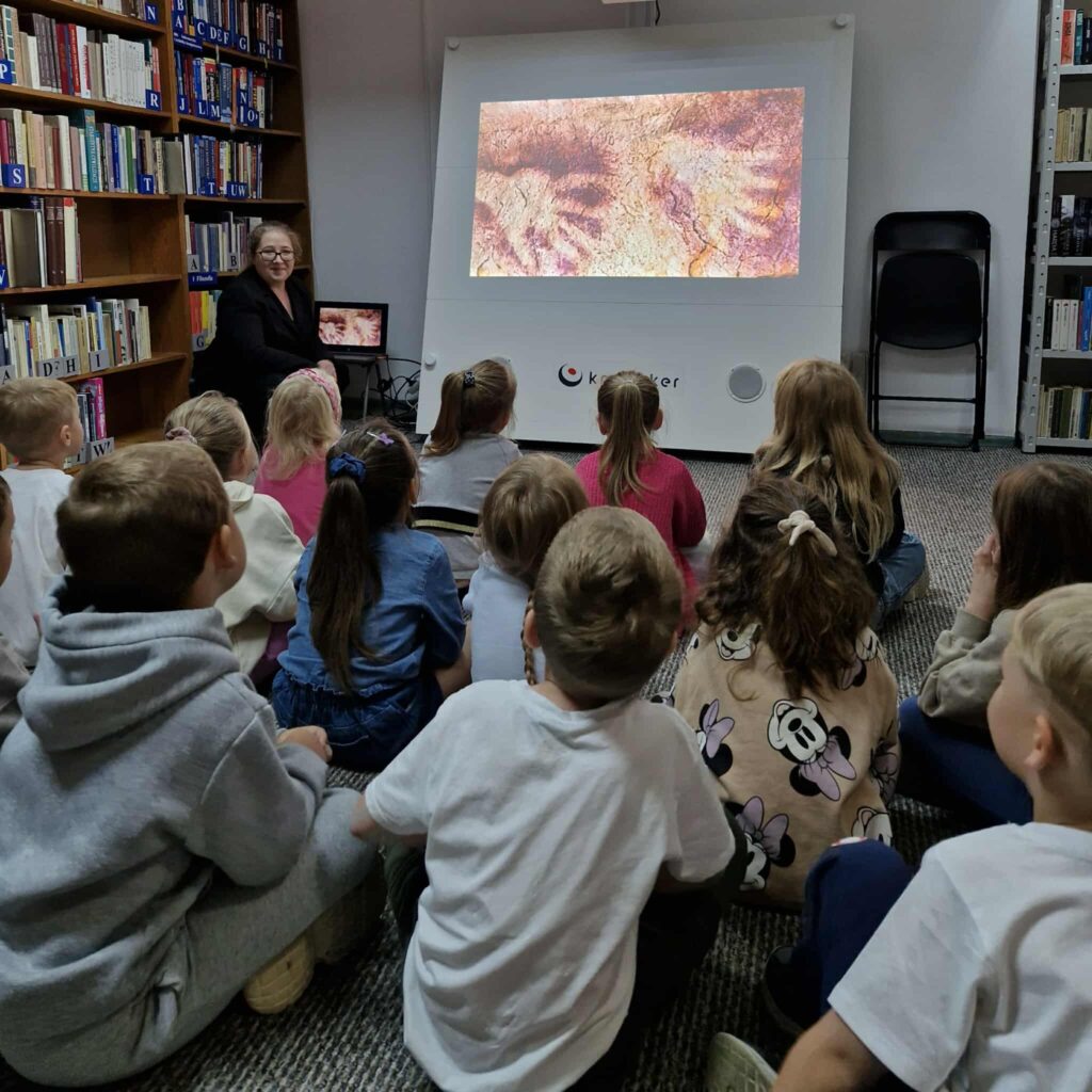 Na zdjęciu widzimy grupę dzieci siedzących na podłodze w bibliotece i oglądających prezentację na ekranie.

Na rzutniku wyświetlany jest obraz przedstawiający malowidła naskalne – zapewne sceny związane ze sztuką prehistoryczną. Obok ekranu siedzi bibliotekarka lub prowadząca zajęcia, która z uśmiechem obserwuje dzieci.

Po lewej stronie widać regały z książkami, co podkreśla biblioteczny charakter miejsca. Dzieci siedzą w skupieniu, zwrócone w stronę ekranu, wyraźnie zainteresowane tematem.

Cała scena przedstawia edukacyjne zajęcia dla najmłodszych w bibliotece, które w ciekawy sposób łączą historię, sztukę i naukę z multimedialną prezentacją.
