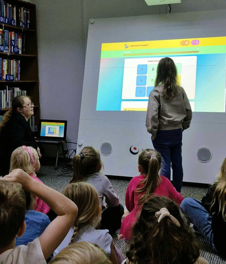 Na zdjęciu grupa dzieci siedzi na dywanie w bibliotece, patrząc na duży interaktywny ekran. Jedno dziecko, odwrócone plecami, stoi przed ekranem i wchodzi z nim w interakcję, podczas gdy reszta obserwuje. W tle widać regały z książkami. Bibliotekarka siedzi z boku i nadzoruje zajęcia.