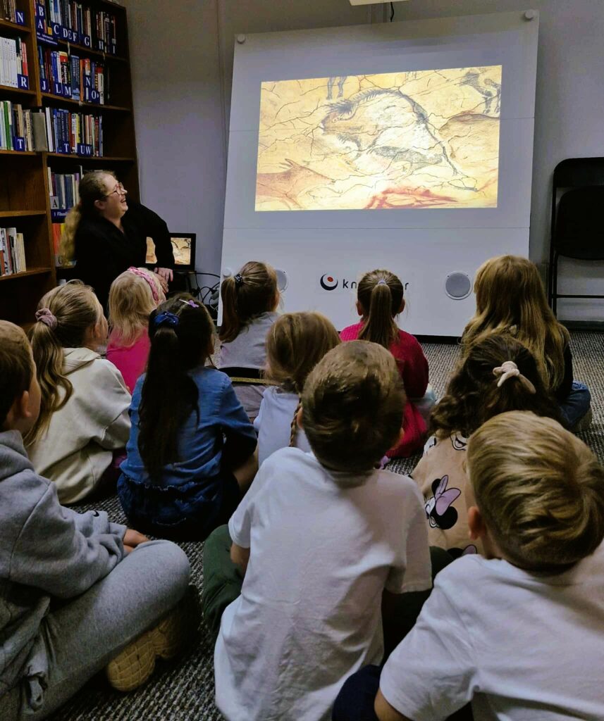Na zdjęciu widzimy grupę dzieci siedzących na podłodze w bibliotece i oglądających prezentację na ekranie.

Na rzutniku wyświetlany jest obraz przedstawiający malowidła naskalne – zapewne sceny związane ze sztuką prehistoryczną. Obok ekranu siedzi bibliotekarka lub prowadząca zajęcia, która z uśmiechem obserwuje dzieci.

Po lewej stronie widać regały z książkami, co podkreśla biblioteczny charakter miejsca. Dzieci siedzą w skupieniu, zwrócone w stronę ekranu, wyraźnie zainteresowane tematem.

Cała scena przedstawia edukacyjne zajęcia dla najmłodszych w bibliotece, które w ciekawy sposób łączą historię, sztukę i naukę z multimedialną prezentacją.