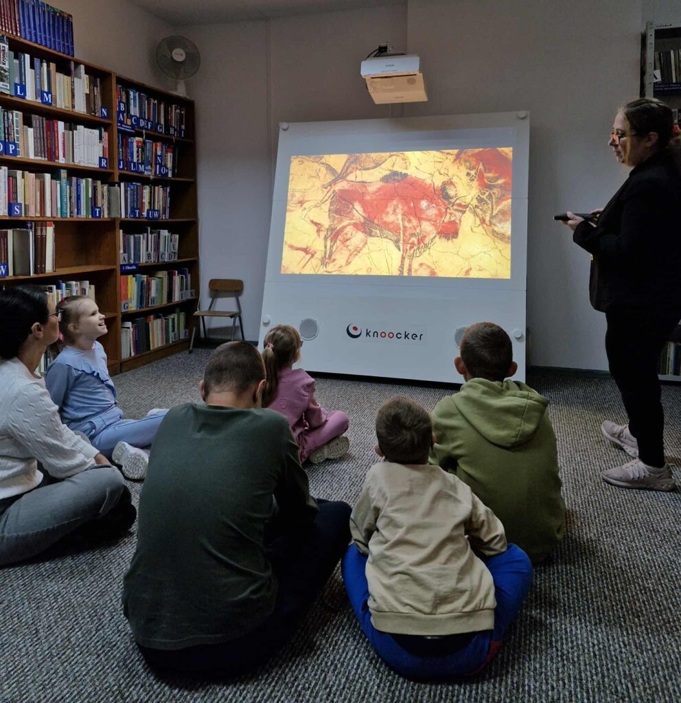 Na zdjęciu widzimy zajęcia edukacyjne prowadzone w bibliotece.
Grupa dzieci siedzi na dywanie i z zainteresowaniem ogląda obraz wyświetlany na interaktywnej tablicy Knoocker. Obok ekranu stoi prowadząca, która obsługuje urządzenie. Na ekranie wyświetlona jest ilustracja przypominająca malowidła naskalne – rysunki wykonane w jaskini, co wskazuje, że tematem zajęć mogą być początki sztuki, historia lub życie ludzi pierwotnych.
W tle widoczne są regały wypełnione książkami, co podkreśla bibliotekarskie otoczenie spotkania. Atmosfera jest skupiona, dzieci są zainteresowane przekazywaną treścią.