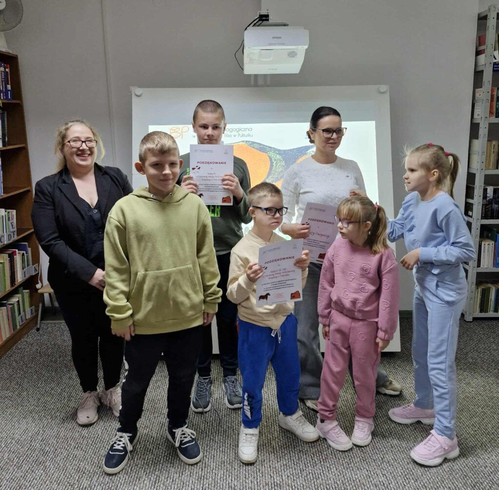 Na zdjęciu widzimy grupę dzieci wraz z bibliotekarką i opiekunką stojących na tle tablicy multimedialnej w bibliotece. Kilkoro dzieci trzyma w dłoniach dyplomy z podziękowaniami za udział w zajęciach. Uczestnicy są uśmiechnięci i dumni ze swoich osiągnięć, a atmosfera jest radosna i pełna satysfakcji.