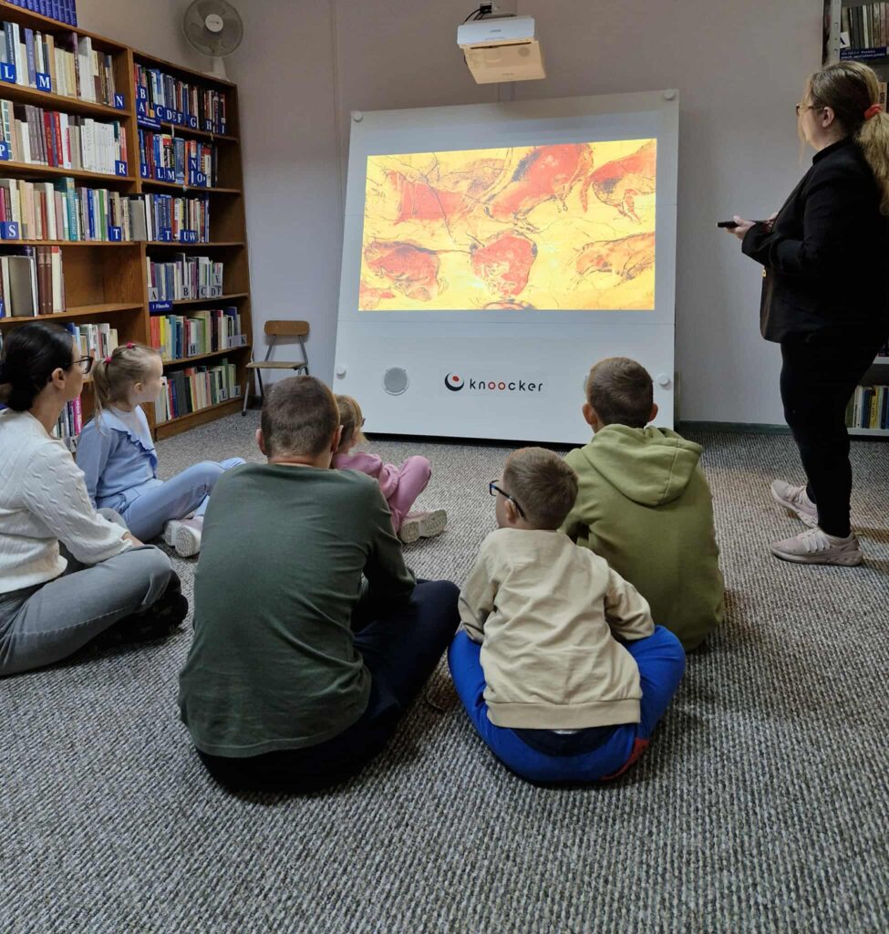 Dzieci i dwie osoby dorosłe siedzą na wykładzinie w bibliotece, skupione na prezentacji wyświetlanej na ekranie systemu „Knoocker”. Na ekranie widoczne są prehistoryczne malowidła jaskiniowe przedstawiające zwierzęta w odcieniach czerwieni i brązu. Jedna z dorosłych osób obsługuje prezentację, druga siedzi wśród dzieci. W tle znajdują się regały pełne książek, co podkreśla edukacyjny charakter miejsca.