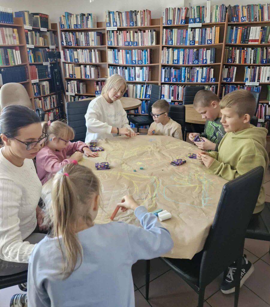 Zdjęcie przedstawia scenę z biblioteki podczas warsztatów kreatywnych. Przy dużym, okrągłym stole pokrytym brązowym papierem pakowym siedzi grupa dzieci oraz trzy osoby dorosłe. Tło wypełniają regały z książkami, uporządkowane alfabetycznie i oznaczone niebieskimi literami od „A” do „AB”. Książki są ustawione pionowo, grzbietami na zewnątrz, a regały wykonane są z jasnego drewna. Pomieszczenie jest dobrze oświetlone naturalnym światłem, które tworzy miękkie cienie na twarzach i przedmiotach.

Po lewej stronie siedzi kobieta o ciemnych włosach spiętych w niski kucyk, w kremowym swetrze o splocie warkoczowym. Ma okulary w czarnej oprawie i z uwagą przygląda się pracy dziecka. Przed nią siedzi dziewczynka w lawendowej bluzie, z blond włosami związanymi w wysoką kitkę różową gumką. Trzyma pomarańczową kredę i rysuje na papierze.
W centrum zdjęcia znajduje się kobieta o jasnych włosach i okrągłych okularach, ubrana w białą bluzę z napisem „Infinite possibilities”. Patrzy spokojnie na dzieci, z rękami opartymi o stół. Przed nią siedzi dziewczynka w różowej bluzie i różowych okularach, rysująca fioletową kredą. Na stole leży fioletowe pudełko z kredą i ilustracjami owoców.
Po prawej stronie siedzi chłopiec w beżowej bluzie, trzymający niebieską kredę, oraz chłopiec w limonkowej bluzie z napisem „GAP”, rysujący fioletową kredą. Na samym końcu siedzi chłopiec w oliwkowej bluzie z krótkimi włosami, który przygląda się trzymanej przez siebie kredzie.
Na papierze widoczne są kolorowe linie i kształty wykonane kredą w odcieniach niebieskiego, żółtego i pomarańczowego. Rozrzucone są także pudełka z kredą oraz pojedyncze kawałki w różnych kolorach. Uczestnicy siedzą na czarnych, skórzanych krzesłach.
Całość ukazuje moment skupionej, twórczej pracy w przyjaznym, edukacyjnym środowisku, gdzie dzieci rozwijają wyobraźnię i zdolności manualne pod opieką dorosłych.