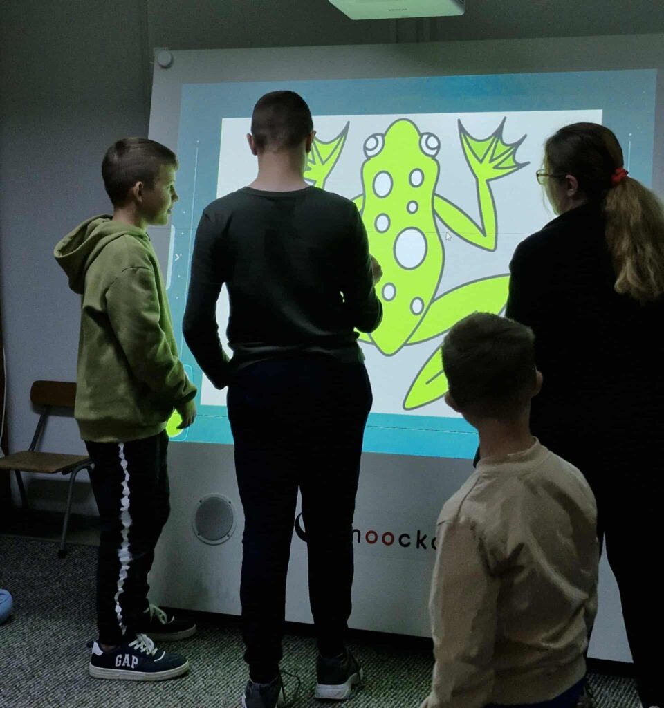Na zdjęciu widać dzieci pracujące przy tablicy multimedialnej w bibliotece. Na ekranie wyświetlona jest duża ilustracja zielonej żaby z białymi plamami. Uczestnicy są skupieni na zadaniu, a obok nich stoi bibliotekarka, która wspiera ich w pracy. Atmosfera sugeruje zajęcia edukacyjne lub interaktywne warsztaty, które łączą naukę z zabawą.