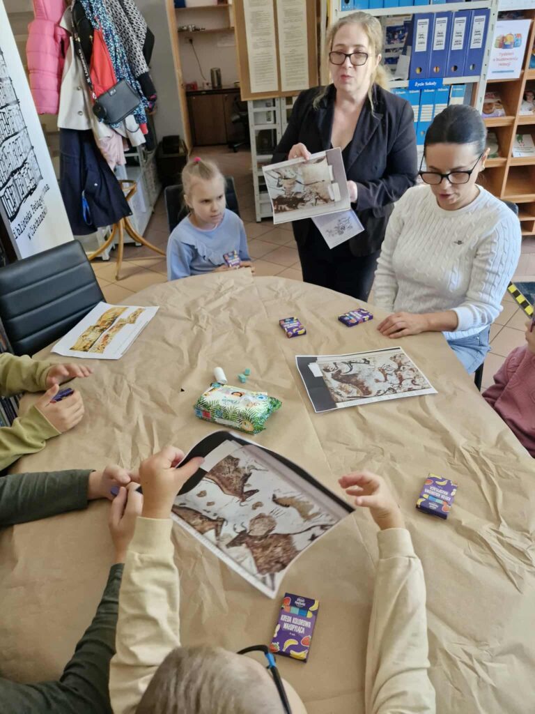 Na zdjęciu widać zajęcia plastyczno-edukacyjne prowadzone w bibliotece.
Przy stole nakrytym papierem siedzi grupa dzieci wraz z osobą dorosłą. Prowadząca zajęcia, stojąca obok, pokazuje kartkę z ilustracją przedstawiającą malowidła naskalne – motyw związany z tematyką prehistorii i sztuki jaskiniowej. Dzieci również trzymają w rękach wydrukowane obrazy, które będą prawdopodobnie stanowić inspirację do ich własnych prac plastycznych.

Na stole leżą kredki oraz kartki, co wskazuje, że uczestnicy przygotowują się do części twórczej warsztatów. Atmosfera jest spokojna i skupiona – dzieci z zainteresowaniem przyglądają się materiałom i słuchają prowadzącej.