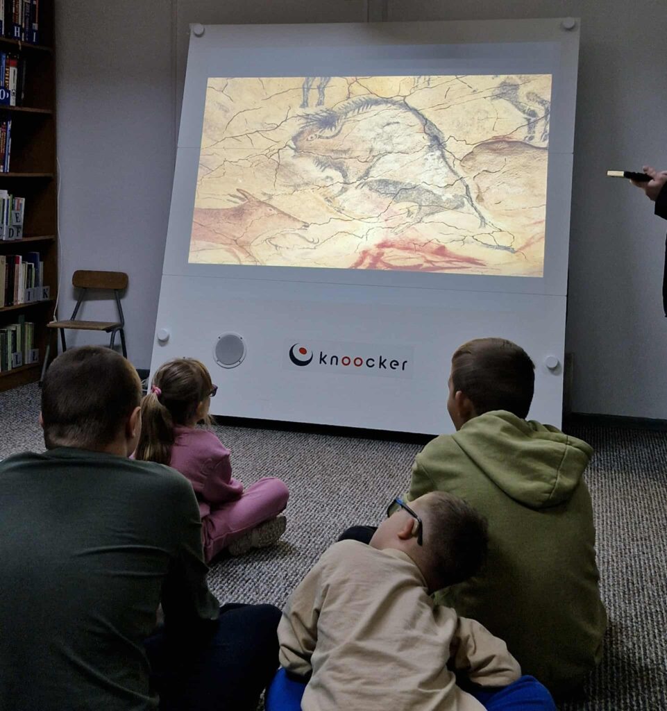 Na zdjęciu widzimy zajęcia edukacyjne prowadzone w bibliotece.
Grupa dzieci siedzi na dywanie i z zainteresowaniem ogląda obraz wyświetlany na interaktywnej tablicy Knoocker. Na ekranie wyświetlona jest ilustracja przypominająca malowidła naskalne – rysunki wykonane w jaskini, co wskazuje, że tematem zajęć mogą być początki sztuki, historia lub życie ludzi pierwotnych.
W tle widoczne są regały wypełnione książkami, co podkreśla bibliotekarskie otoczenie spotkania. Atmosfera jest skupiona, dzieci są zainteresowane przekazywaną treścią.