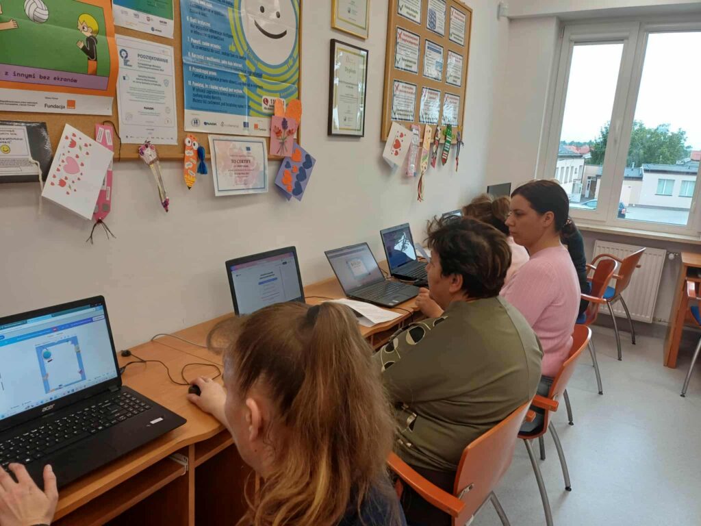 W sali komputerowej pięć osób siedzi przy biurkach, pracując na laptopach. Na ekranach widoczne są strony internetowe i formularze, co sugeruje udział w szkoleniu lub kursie cyfrowym. Ściany ozdobione są certyfikatami, plakatami edukacyjnymi i dziecięcymi pracami plastycznymi. Duże okna wpuszczają naturalne światło, tworząc przyjazną atmosferę nauki.