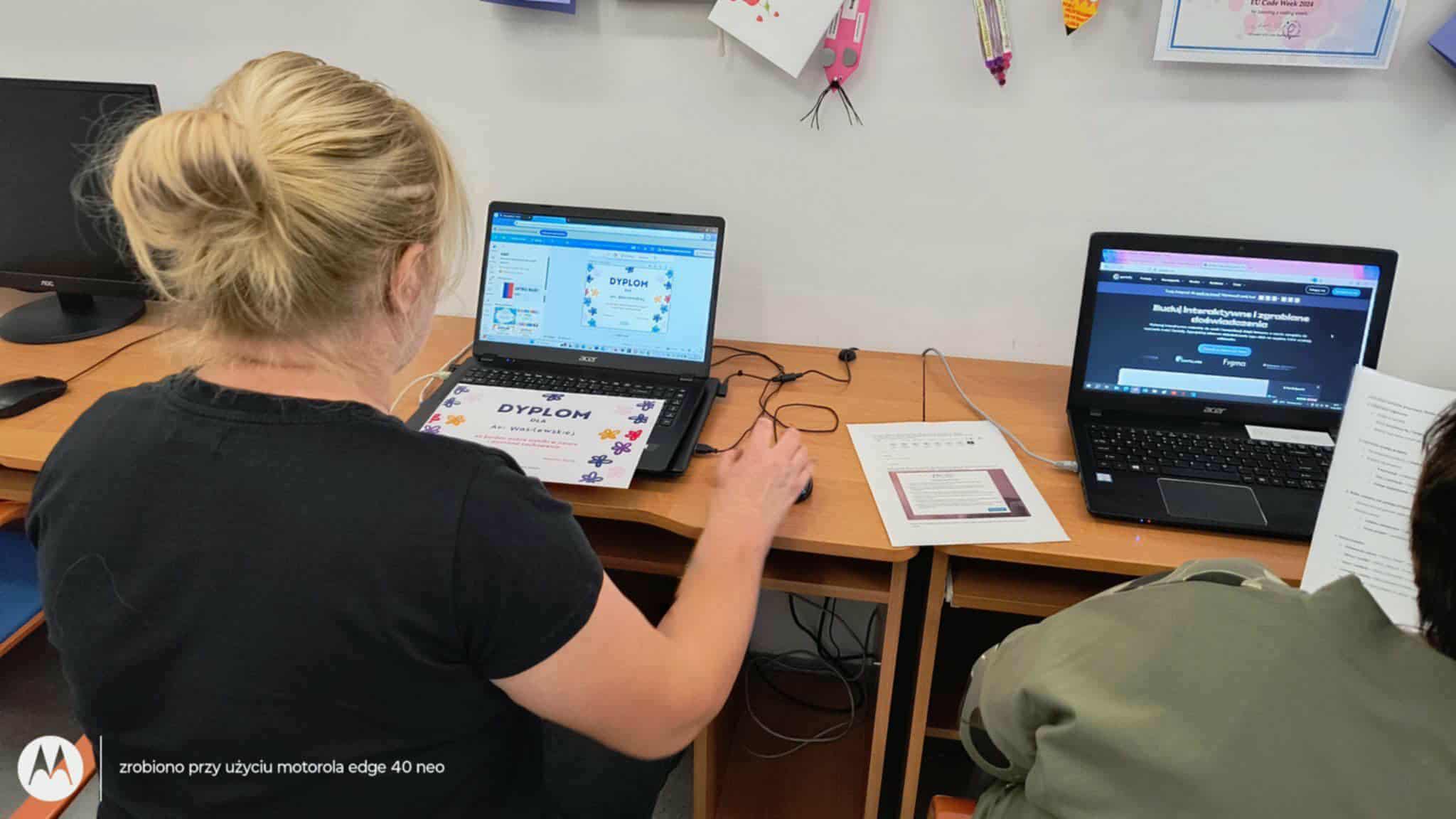 Dwie osoby siedzą przy biurku i pracują na laptopach. Na jednym z ekranów wyświetlony jest dyplom z grafiką puzzli, a jego papierowa wersja leży na klawiaturze. Drugi laptop pokazuje stronę internetową z tekstem w języku polskim. Między komputerami znajdują się dodatkowe materiały. W tle widać dziecięce prace plastyczne na ścianie, co sugeruje edukacyjne otoczenie.