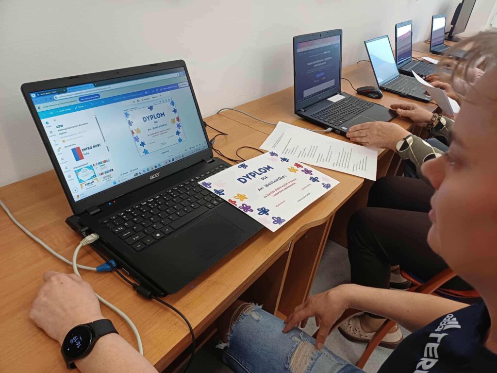 W sali komputerowej uczestnicy pracują na laptopach, skupieni na zadaniach edukacyjnych. Na ekranie jednego z komputerów widoczny jest kolorowy dyplom tworzony w Canvie, a jego papierowa wersja leży na biurku. W tle widać inne osoby pracujące przy stanowiskach oraz dodatkowe materiały. Całość ukazuje atmosferę nauki i kreatywności w przyjaznym, edukacyjnym otoczeniu.