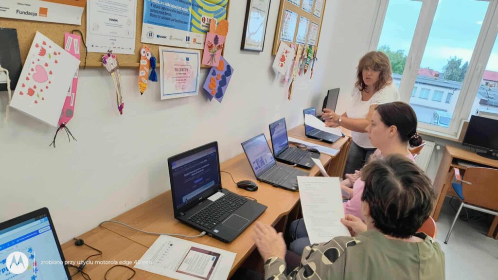 W sali szkoleniowej uczestnicy siedzą przy biurkach z laptopami, zaangażowani w naukę z zakresu kompetencji cyfrowych. Prowadząca osoba stoi i udziela wskazówek. Na ścianach widoczne są kolorowe dekoracje, plakaty informacyjne i certyfikaty, co nadaje przestrzeni charakter edukacyjny i wspólnotowy. Całość sugeruje warsztaty dla dorosłych w przyjaznym, lokalnym środowisku.