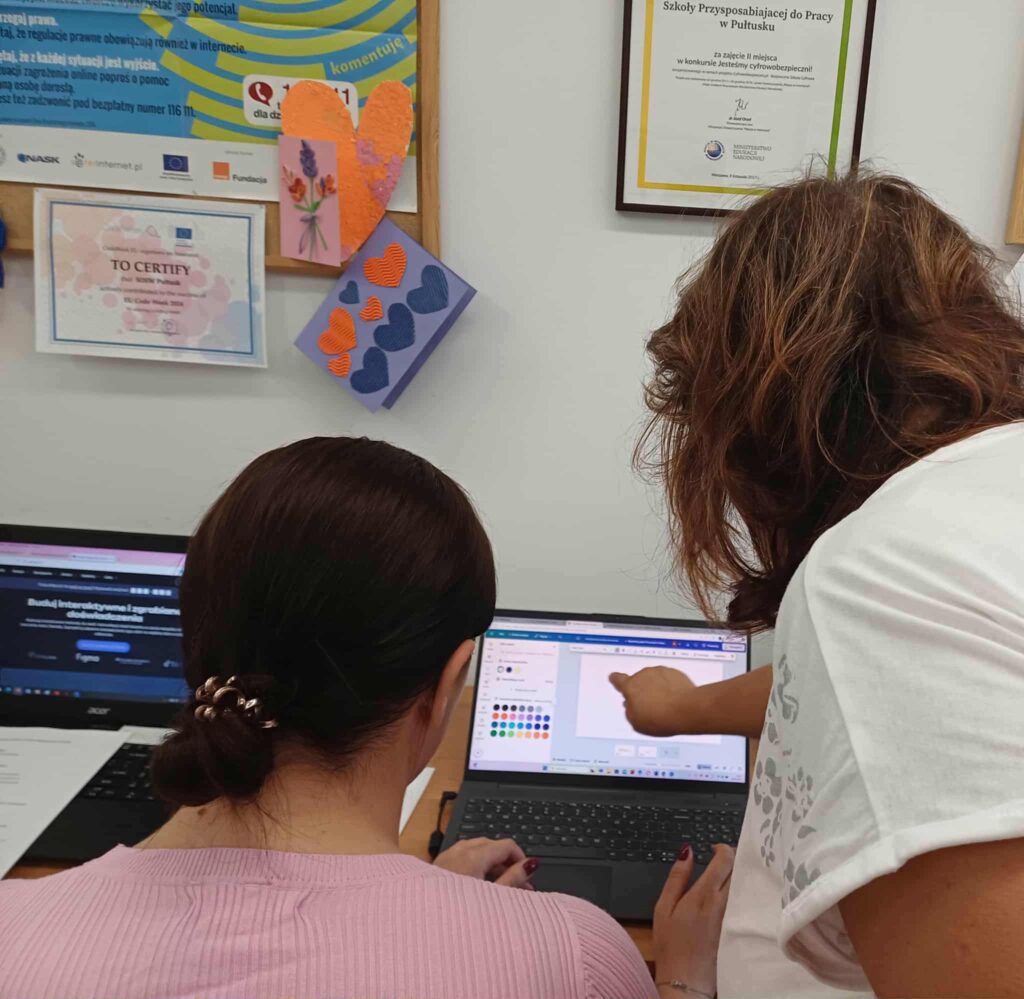 Dwie kobiety pracują wspólnie przy biurku z laptopami w przestrzeni edukacyjnej. Jedna z nich siedzi w różowej bluzce, z włosami spiętymi ozdobną gumką, druga stoi obok i wskazuje coś na ekranie — prawdopodobnie w programie graficznym z paletą kolorów. W tle widoczne są certyfikaty, plakaty i kolorowe papierowe serca, co nadaje miejscu kreatywny i szkoleniowy charakter. Całość ukazuje atmosferę współpracy i nauki w przyjaznym otoczeniu.