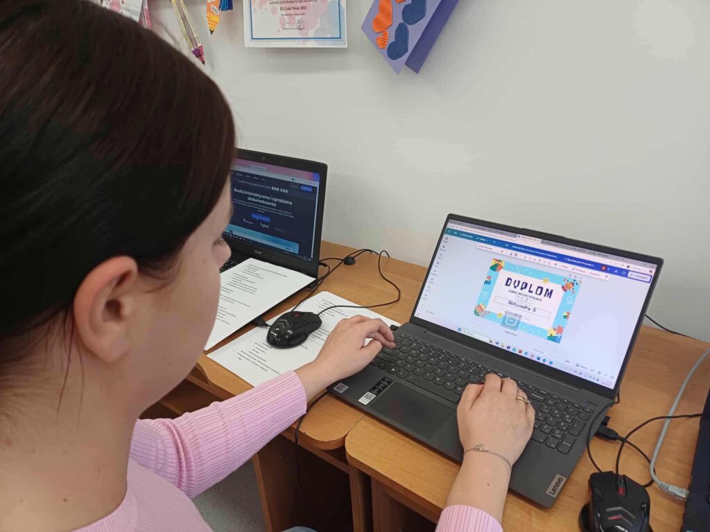 Kobieta w różowej bluzce pracuje przy biurku na dwóch laptopach. Na jednym z ekranów edytuje kolorowy dyplom w Canvie, a obok znajduje się strona internetowa. Na ścianie w tle wiszą certyfikaty i dekoracje z papierowymi sercami, co nadaje przestrzeni edukacyjny i kreatywny charakter.