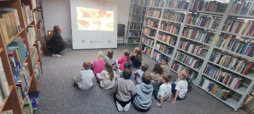 Na zdjęciu widzimy grupę dzieci siedzących na podłodze w bibliotece i oglądających prezentację na ekranie.

Na rzutniku wyświetlany jest obraz przedstawiający malowidła naskalne – zapewne sceny związane ze sztuką prehistoryczną. Obok ekranu siedzi bibliotekarka lub prowadząca zajęcia, która z uśmiechem obserwuje dzieci.

Po lewej stronie widać regały z książkami, co podkreśla biblioteczny charakter miejsca. Dzieci siedzą w skupieniu, zwrócone w stronę ekranu, wyraźnie zainteresowane tematem.

Cała scena przedstawia edukacyjne zajęcia dla najmłodszych w bibliotece, które w ciekawy sposób łączą historię, sztukę i naukę z multimedialną prezentacją.