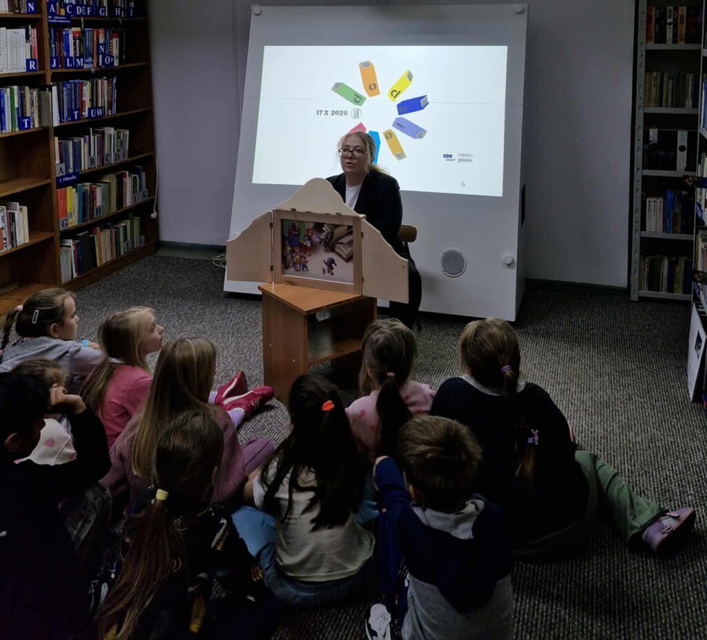 Na zdjęciu widać bibliotekarkę prowadzącą zajęcia dla grupy dzieci w bibliotece. Kobieta siedzi przy drewnianym teatrzyku kamishibai, w którym widoczna jest ilustracja do opowieści. Dzieci siedzą na dywanie, skupione i zasłuchane w historię.
W tle znajduje się ekran z wyświetlonym kolorowym logotypem akcji „Noc Bibliotek 2025” oraz datą 17 X 2025. Na półkach po bokach widać książki starannie ułożone i oznaczone literami.
Zdjęcie oddaje atmosferę ciepła, skupienia i zaciekawienia – to moment wspólnego czytania, który promuje ideę biblioteki jako miejsca spotkania z literaturą i kulturą.
