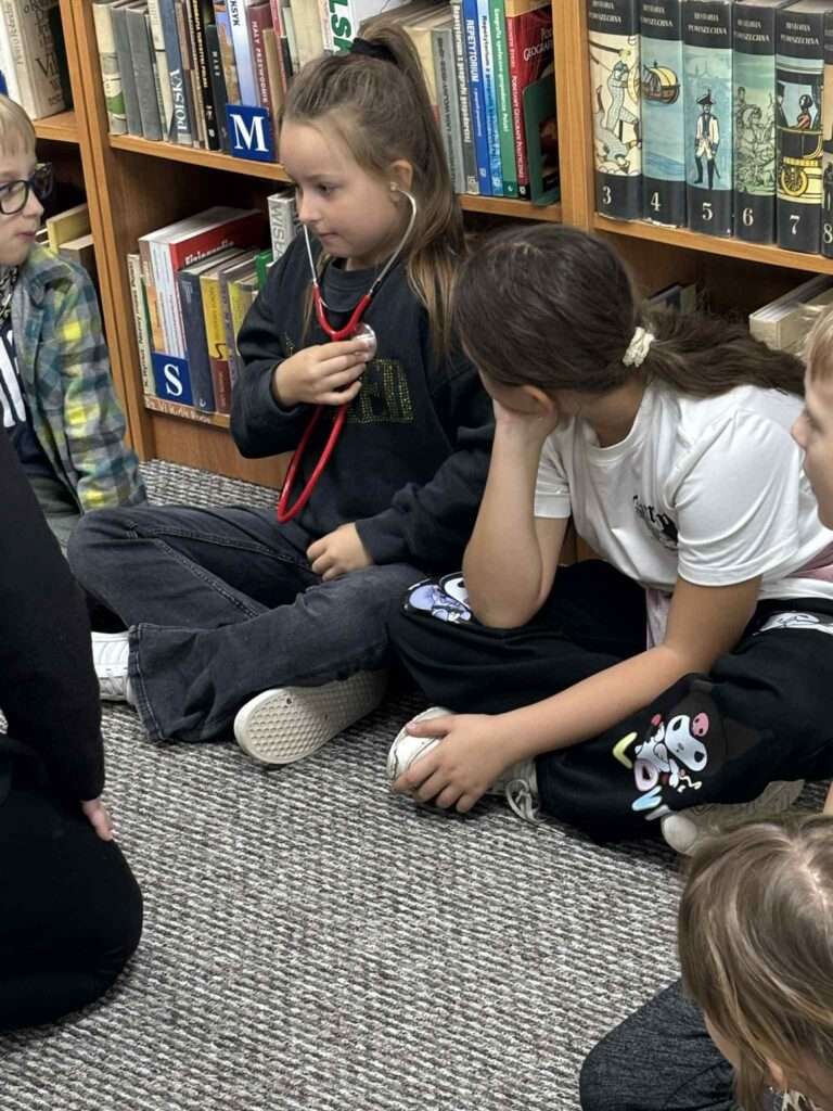 Na zdjęciu widać grupę dzieci siedzących na dywanie w bibliotece. Jedna z dziewczynek trzyma stetoskop przy klatce piersiowej i przysłuchuje się biciu własnego serca, a reszta dzieci z zaciekawieniem obserwuje to doświadczenie. W tle widoczne są regały z książkami. Zdjęcie oddaje atmosferę zainteresowania, nauki i wspólnej zabawy podczas zajęć w bibliotece.