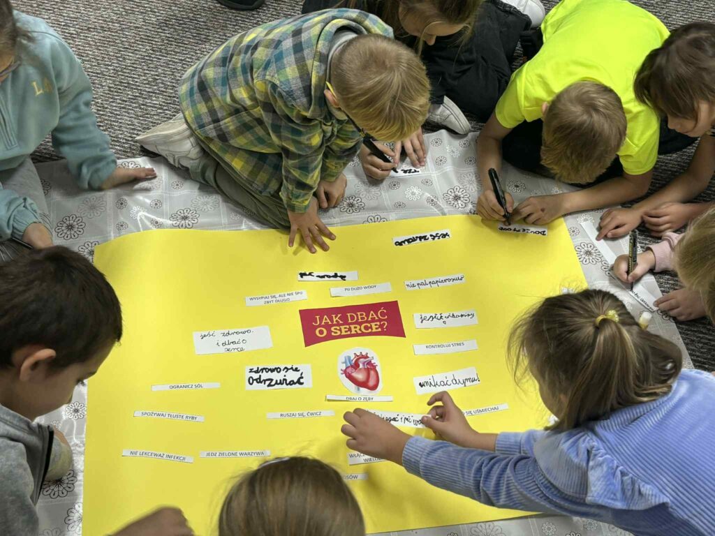 Grupa dzieci siedzi wokół dużego żółtego plakatu z napisem „JAK DBAĆ O SERCE?” i ilustracją serca. Dzieci aktywnie uczestniczą w zadaniu — piszą na małych kartkach i przyklejają je do plakatu, dzieląc się pomysłami na temat dbania o zdrowie serca. Całość ukazuje angażującą, zdrowotną aktywność edukacyjną w duchu współpracy i kreatywności.