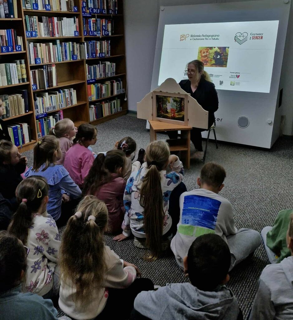 Na zdjęciu widać spotkanie z dziećmi w bibliotece, podczas którego odbywa się głośne czytanie w ramach akcji „Czytamy z sercem”.
Na tle regałów z książkami i ekranu multimedialnego z logotypami akcji siedzi bibliotekarka, która prowadzi pokaz teatrzyku kamishibai – drewnianego teatrzyku obrazkowego. Wewnątrz teatrzyku widoczna jest ilustracja przedstawiająca scenę z bajki, której słuchają dzieci.
Przed bibliotekarką, na dywanie, siedzi grupa uważnych słuchaczy – dzieci, skupione i zaciekawione opowieścią. Cała scena ma ciepły, kameralny charakter i ukazuje bibliotekę jako miejsce przyjazne młodym czytelnikom, promujące kulturę czytania i rozwijanie empatii.