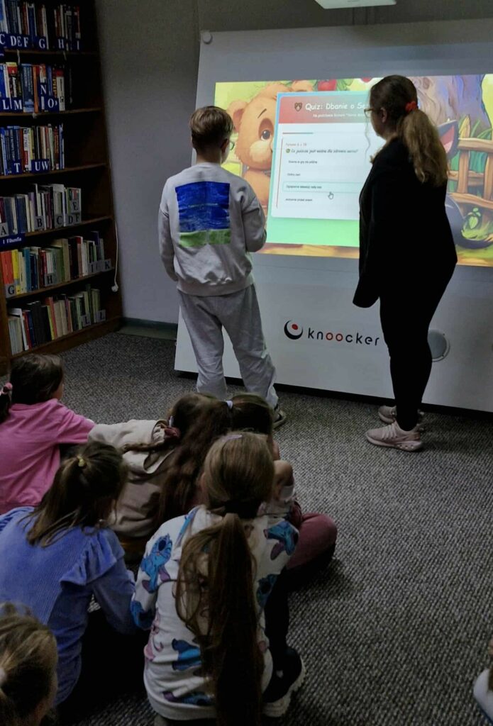 Na zdjęciu widać grupę dzieci uczestniczących w zajęciach w bibliotece, realizowanych w ramach akcji „Czytamy z sercem”. Uczestnicy siedzą na podłodze, z zainteresowaniem obserwując duży interaktywny ekran, na którym wyświetlony jest quiz o zdrowiu serca.
Jedno z dzieci odpowiada na pytania, korzystając z dotykowego ekranu, a obok stoi prowadząca zajęcia bibliotekarka, która czuwa nad przebiegiem zabawy. Na ekranie widać pytanie: „Co jest dobre dla zdrowia serca?”, a tło przedstawia ilustrację z sympatycznym misiem — element nawiązujący do wcześniejszego teatrzyku kamishibai.
Spotkanie łączy edukację zdrowotną z promocją czytelnictwa i nowoczesnymi technologiami, tworząc atmosferę nauki przez zabawę w przyjaznej przestrzeni biblioteki.