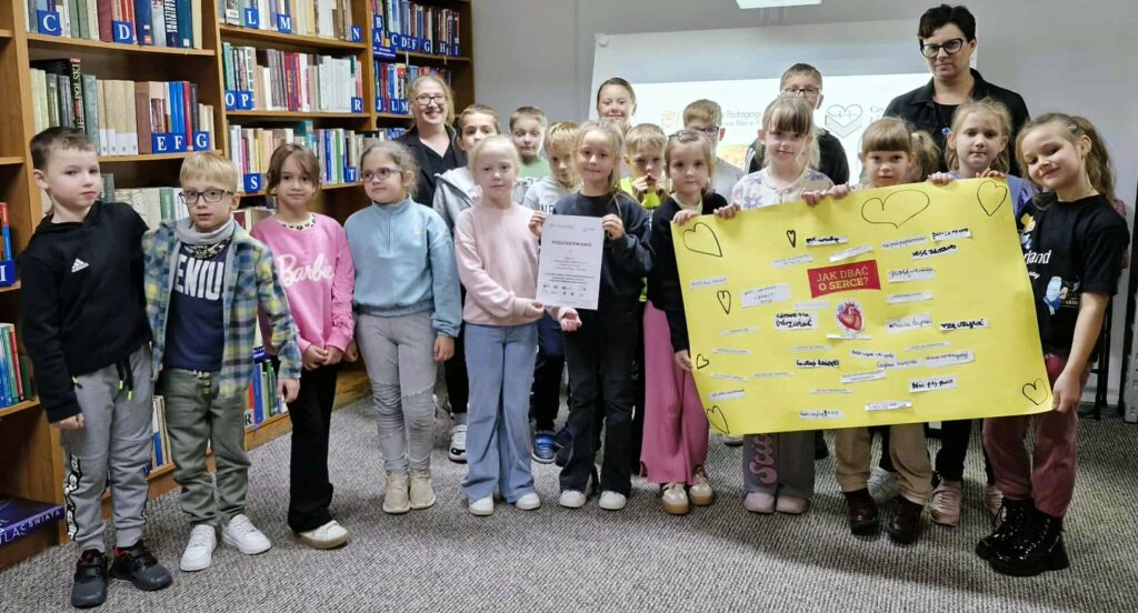 Na zdjęciu widoczna jest duża grupa dzieci oraz dwie dorosłe kobiety stojące w bibliotece, przed regałami z książkami. Dzieci trzymają duży, żółty plakat z tytułem "JAK DBAĆ O SERCE?" oraz notatkami i rysunkiem serca. Część dzieci trzyma również certyfikaty lub dyplomy. Wszyscy uśmiechają się do zdjęcia, prezentując efekty swojej pracy.
