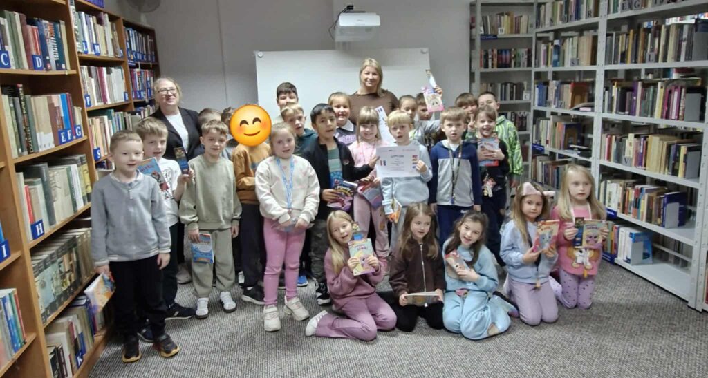 Na zdjęciu widać grupę dzieci wraz z dwiema dorosłymi osobami w bibliotece. Uczestnicy stoją i siedzą wśród regałów pełnych książek, uśmiechają się i trzymają w rękach różne książki lub dyplomy. Na twarzach dzieci widać radość i zainteresowanie — atmosfera jest przyjazna i pełna entuzjazmu.
Zdjęcie zostało wykonane podczas zajęć bibliotecznych promujących czytelnictwo w ramach akcji „Noc Bibliotek”. W tle widać uporządkowane regały z książkami oznaczonymi literami alfabetu, a na ścianie – biały ekran projekcyjny.
Całość oddaje ciepły, edukacyjny klimat biblioteki jako miejsca wspólnej zabawy i nauki.