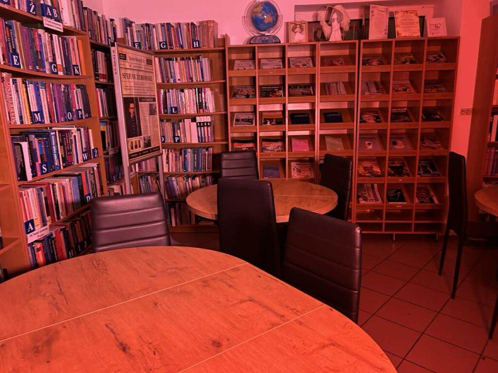 Na zdjęciu widać wnętrze biblioteki czytelni, o przytulnym, kameralnym charakterze. Pomieszczenie jest oświetlone intensywnym, czerwonym światłem, które nadaje całości nietypowy, niemal sceniczny klimat.

Co znajduje się na zdjęciu?

Stoliki i krzesła
Przednie plany zajmują okrągłe, drewniane stoły oraz czarne, tapicerowane krzesła. Wszystko wygląda przygotowane do pracy w małych grupach, cichego czytania lub spotkań.

Regały z książkami
Z lewej strony widoczne są wysokie regały wypełnione książkami, posegregowanymi alfabetycznie (widoczne literowe etykiety: A, B, E, G, K, M, O, P, S, W, Ż). Książki stoją gęsto, co wskazuje na bogaty księgozbiór.

Gazetki i czasopisma
Na prawym regale znajdują się otwarte półki z czasopismami i materiałami edukacyjnymi ułożonymi warstwowo.

Plansza z informacją / wystawa
W centralnej części, między regałami, stoi duża plansza ekspozycyjna – prawdopodobnie część jakiejś wystawy tematycznej. Widać na niej fragment tekstu i fotografię osoby historycznej.

Dekoracje i ekspozycje
Na górze środkowego regału stoją dodatkowe elementy dekoracyjne: globus, figurki lub prace plastyczne oraz otwarte książki lub albumy eksponowane na stojakach.

Ogólne wrażenie

To ciche, uporządkowane miejsce sprzyjające nauce i czytaniu. Czerwone światło może być wynikiem ustawień aparatu lub oświetlenia, co tworzy niecodzienny efekt wizualny, ale sama przestrzeń prezentuje się funkcjonalnie i estetycznie.