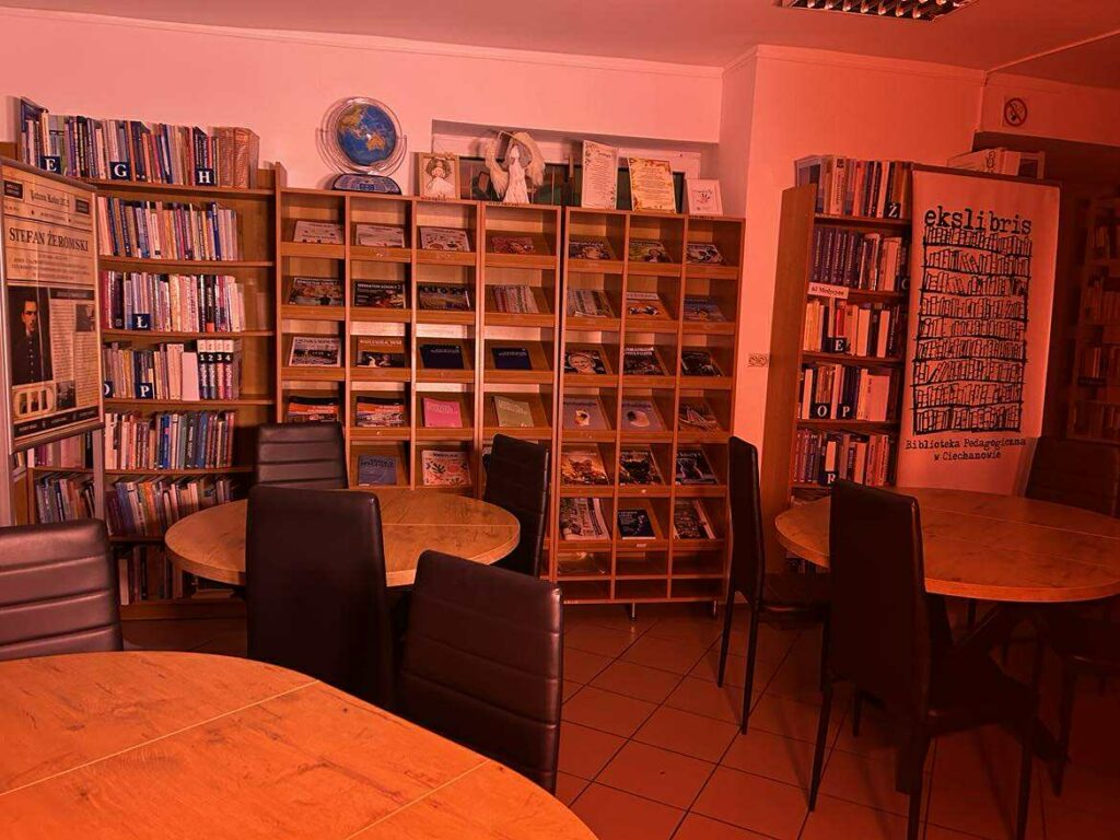 Na zdjęciu widać wnętrze biblioteki czytelni, o przytulnym, kameralnym charakterze. Pomieszczenie jest oświetlone intensywnym, czerwonym światłem, które nadaje całości nietypowy, niemal sceniczny klimat.

Co znajduje się na zdjęciu?

Stoliki i krzesła
Przednie plany zajmują okrągłe, drewniane stoły oraz czarne, tapicerowane krzesła. Wszystko wygląda przygotowane do pracy w małych grupach, cichego czytania lub spotkań.

Regały z książkami
Z lewej strony widoczne są wysokie regały wypełnione książkami, posegregowanymi alfabetycznie (widoczne literowe etykiety: A, B, E, G, K, M, O, P, S, W, Ż). Książki stoją gęsto, co wskazuje na bogaty księgozbiór.

Gazetki i czasopisma
Na prawym regale znajdują się otwarte półki z czasopismami i materiałami edukacyjnymi ułożonymi warstwowo.

Plansza z informacją / wystawa
W centralnej części, między regałami, stoi duża plansza ekspozycyjna – prawdopodobnie część jakiejś wystawy tematycznej. Widać na niej fragment tekstu i fotografię osoby historycznej.

Dekoracje i ekspozycje
Na górze środkowego regału stoją dodatkowe elementy dekoracyjne: globus, figurki lub prace plastyczne oraz otwarte książki lub albumy eksponowane na stojakach.

Ogólne wrażenie

To ciche, uporządkowane miejsce sprzyjające nauce i czytaniu. Czerwone światło może być wynikiem ustawień aparatu lub oświetlenia, co tworzy niecodzienny efekt wizualny, ale sama przestrzeń prezentuje się funkcjonalnie i estetycznie.