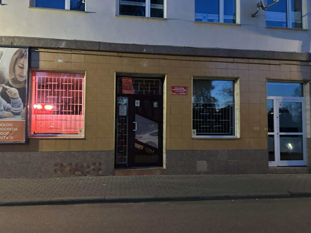 Na zdjęciu widać fasadę budynku, w którym mieści się instytucja publiczna — po lewej stronie znajduje się reklama usług psychologiczno-pedagogicznych, a na środku przy głównym wejściu widoczna jest czerwona tabliczka z napisem wskazującym na bibliotekę pedagogiczną (bez podawania, jaka dokładnie, zgodnie z zasadami).

Co widać na zdjęciu?

Wejście główne
Centralnie znajduje się ciemne drzwi wejściowe z przeszkloną częścią. Nad nimi, po prawej stronie, jest czerwona tabliczka informacyjna instytucji. Drzwi wyglądają na zamknięte, ale oklejone są naklejkami informacyjnymi (np. godziny pracy, zasady wejścia).

Okna z kratami
Po obu stronach drzwi są dwa duże okna zabezpieczone metalowymi kratami.

Lewe okno świeci intensywnym czerwonym światłem — to światło z wnętrza biblioteki, które widziałaś/eś na wcześniejszym zdjęciu.

Prawe okno odbija niebo, drzewa i otoczenie, sugerując, że zdjęcie zostało zrobione o zmierzchu lub wczesnym wieczorem.

Reklama po lewej
Reklama z wizerunkiem dorosłej osoby i dziecka wskazuje, że obok mieści się poradnia lub gabinet specjalistyczny.

Otoczenie
Przed budynkiem jest chodnik z kostki brukowej. Ujęcie wykonano z poziomu ulicy.

Ogólne wrażenie

Budynek wygląda na siedzibę instytucji oświatowej lub usług edukacyjnych. Ciepłe, czerwone światło w lewym oknie wyróżnia się na tle chłodnych barw zmierzchu, tworząc ciekawy efekt wizualny. Całość sprawia wrażenie miejsca zadbanego, spokojnego i uporządkowanego.