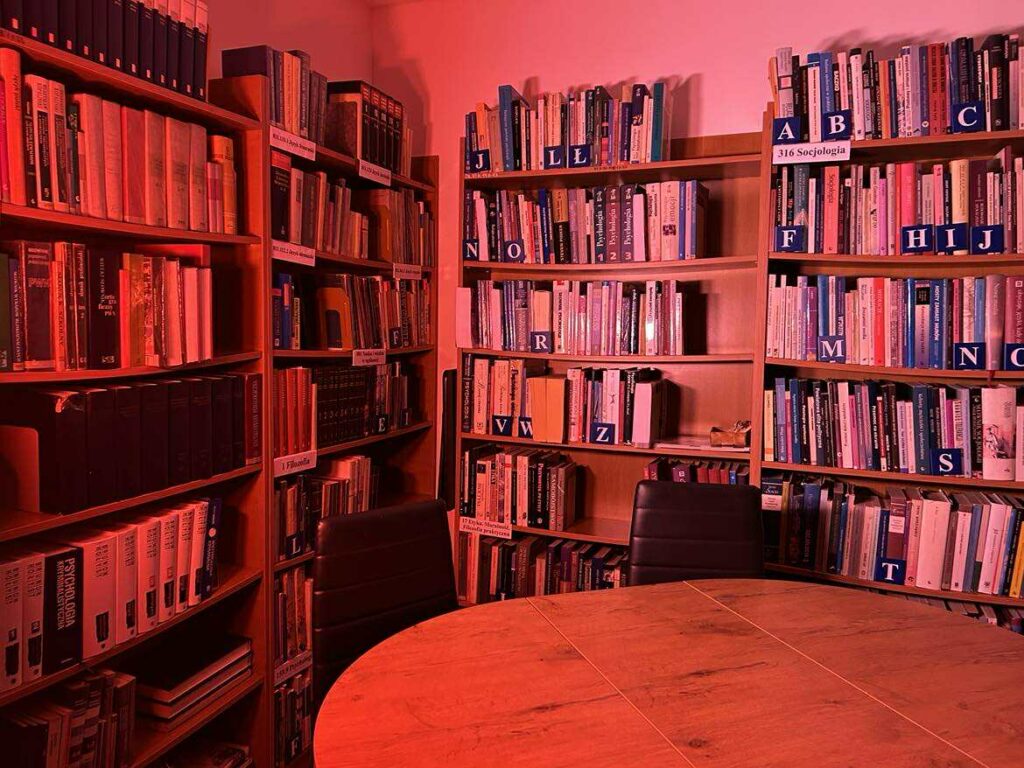 Na zdjęciu widać wnętrze czytelni, o przytulnym, kameralnym charakterze. Pomieszczenie jest oświetlone intensywnym, czerwonym światłem, które nadaje całości nietypowy, niemal sceniczny klimat.

Co znajduje się na zdjęciu?
Regały z książkami
Z lewej strony widoczne są wysokie regały wypełnione książkami, posegregowanymi alfabetycznie (widoczne literowe etykiety: A, B, E, G, K, M, O, P, S, W, Ż). Książki stoją gęsto, co wskazuje na bogaty księgozbiór.
Stoliki i krzesła
Przednie plany zajmują okrągłe, drewniane stoły oraz czarne, tapicerowane krzesła. Wszystko wygląda przygotowane do pracy w małych grupach, cichego czytania lub spotkań.
To ciche, uporządkowane miejsce sprzyjające nauce i czytaniu. Czerwone światło tworzy niecodzienny efekt wizualny, ale sama przestrzeń prezentuje się funkcjonalnie i estetycznie.