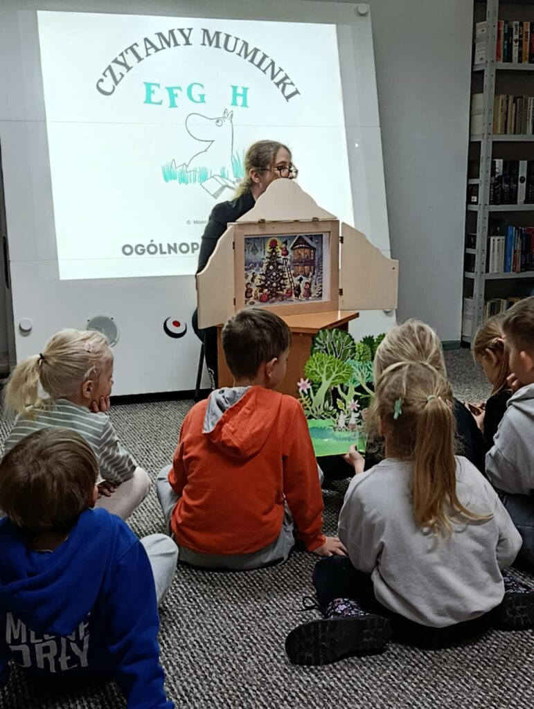 Na zdjęciu widać fragment zajęć czytelniczych prowadzonych dla dzieci w bibliotece w ramach ogólnopolskiej akcji „Czytamy Muminki”. Prowadząca siedzi przy drewnianej skrzynce kamishibai, w której umieszczona jest ilustracja przedstawiająca świąteczną scenę. Przed nią, na dywanie, siedzi grupa dzieci zasłuchanych i skupionych na opowieści. W tle widać tablicę z projekcją logo akcji oraz regały pełne książek. Atmosfera jest ciepła i kameralna – dzieci żywo uczestniczą w spotkaniu i śledzą prezentowaną historię.
