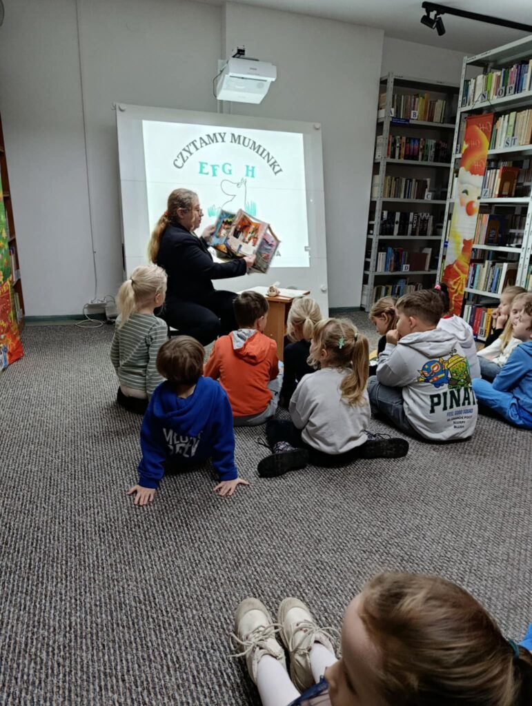 Na zdjęciu widać zajęcia biblioteczne prowadzone dla grupy małych dzieci. W centralnej części siedzi osoba dorosła – prowadząca zajęcia – trzymająca otwartą, kolorową książkę i pokazująca ilustracje dzieciom. Za prowadzącą znajduje się duży ekran lub tablica multimedialna z wyświetlonym logotypem akcji „Czytamy Muminki”, przedstawiającym charakterystyczną kreskę Muminków oraz napis EFGH.
Dzieci siedzą na dywanie w półkolu przed osobą czytającą – większość jest zwrócona w jej stronę i słucha z zainteresowaniem. Są ubrane w kolorowe bluzy i swobodną odzież.
Po prawej stronie fotografii widać półki pełne książek oraz fragment dekoracji z rysunkiem o świątecznym motywie.
Całość tworzy ciepłą, edukacyjną atmosferę wspólnego czytania i bibliotecznej pracy z najmłodszymi.