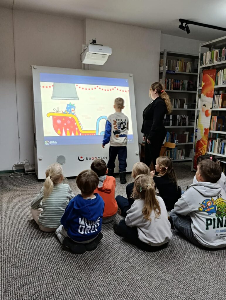 Na zdjęciu widać zajęcia edukacyjne prowadzone dla grupy dzieci w bibliotece. Dzieci siedzą na dywanie przed dużą interaktywną tablicą, na której wyświetlana jest kolorowa, bajkowa animacja. Jedno z dzieci stoi przy ekranie i bierze udział w zadaniu, a obok niego stoi prowadząca zajęcia, obserwując i wspierając uczestnika. W tle widoczne są regały pełne książek, co podkreśla biblioteczny charakter spotkania. Atmosfera jest skupiona, a dzieci wyglądają na zainteresowane prezentowanymi treściami.