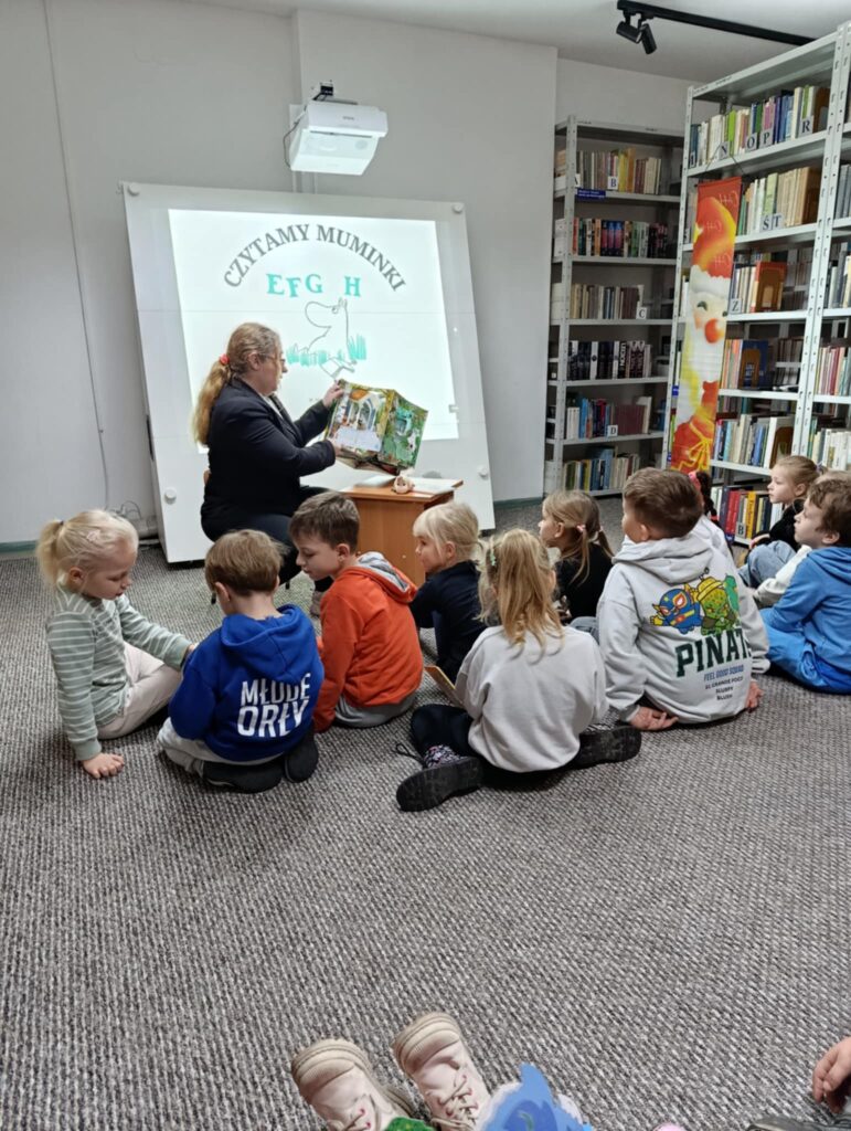 Na zdjęciu widać zajęcia biblioteczne prowadzone dla grupy małych dzieci. W centralnej części siedzi osoba dorosła – prowadząca zajęcia – trzymająca otwartą, kolorową książkę i pokazująca ilustracje dzieciom. Za prowadzącą znajduje się duży ekran lub tablica multimedialna z wyświetlonym logotypem akcji „Czytamy Muminki”, przedstawiającym charakterystyczną kreskę Muminków oraz napis EFGH.
Dzieci siedzą na dywanie w półkolu przed osobą czytającą – większość jest zwrócona w jej stronę i słucha z zainteresowaniem. Są ubrane w kolorowe bluzy i swobodną odzież.
Po prawej stronie fotografii widać półki pełne książek oraz fragment dekoracji z rysunkiem o świątecznym motywie.
Całość tworzy ciepłą, edukacyjną atmosferę wspólnego czytania i bibliotecznej pracy z najmłodszymi.