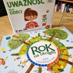 Na zdjęciu widoczne są materiały edukacyjne dla dzieci, ułożone na drewnianym stole w bibliotece.

Na pierwszym planie znajduje się kolorowa gra „Rok na okrągło. Gra obserwacyjna”. Jej okładka przedstawia okrągły schemat roku podzielony na pory roku, wokół którego widoczne są scenki z życia dzieci – zabawy na świeżym powietrzu, jazda na rowerze, lepienie bałwana czy piknik. Ilustracje są żywe, pełne detali i zachęcają do obserwacji oraz rozmowy o zmianach zachodzących w przyrodzie.

Za grą ustawione jest pudełko zestawu edukacyjnego „Trening mindfulness. Uważność dla dzieci” (oznaczone jako 4+). Na opakowaniu widnieje ilustracja dziewczynki oraz elementy przyrody – kwiatek i pszczółka – co sugeruje spokojny, przyjazny charakter ćwiczeń. Podtytuł informuje o 50 zabawach wspierających koncentrację, co wskazuje na rozwijanie uważności i umiejętności skupienia u najmłodszych.

W tle widoczne są regały z książkami, co potwierdza, że zdjęcie zostało wykonane w przestrzeni bibliotecznej lub edukacyjnej. Całość przedstawia atrakcyjne, nowoczesne pomoce dydaktyczne wspierające rozwój poznawczy i emocjonalny dzieci.